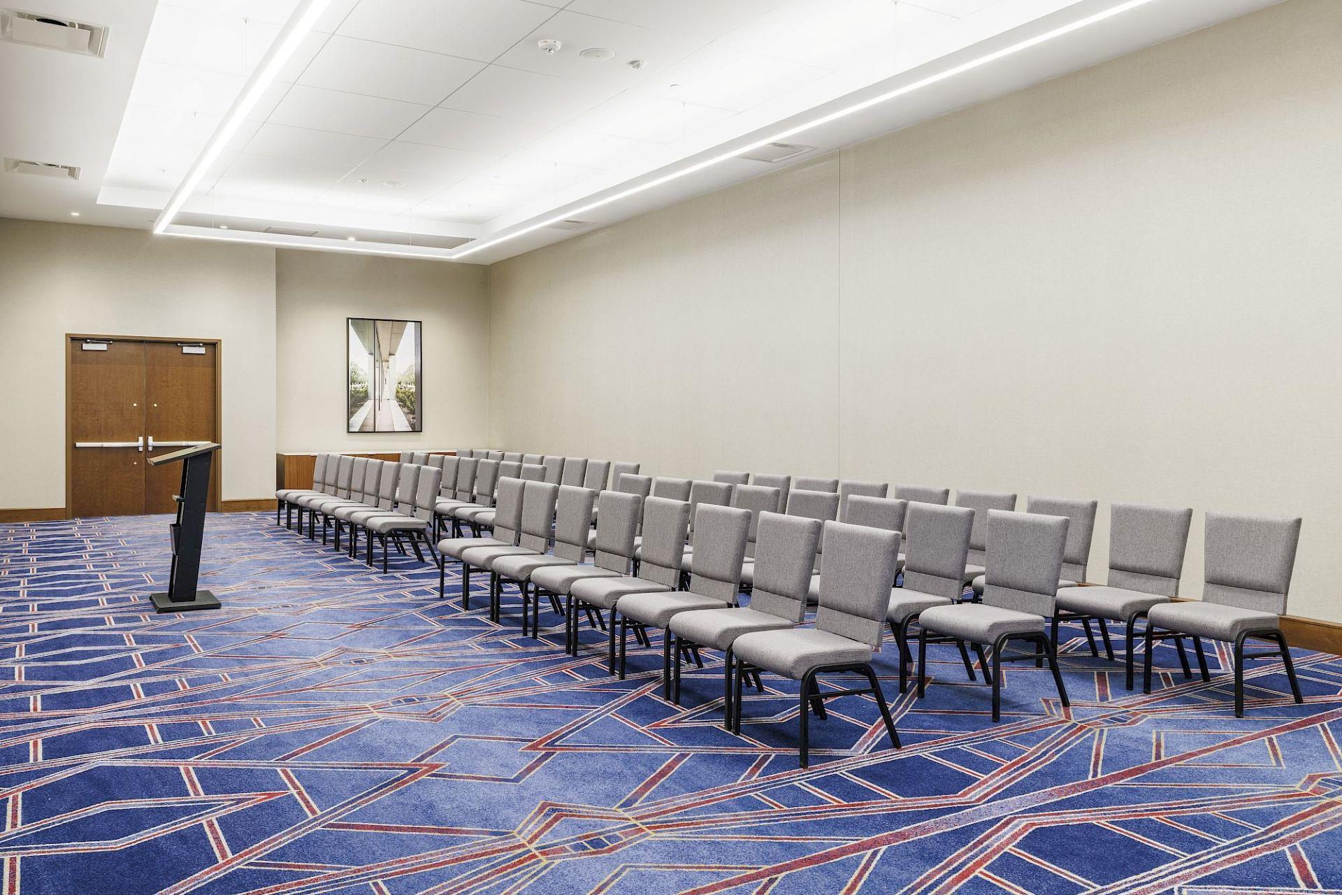 Conference room set theater-style with rows of gray chairs facing a lectern, framed artwork, and blue geometric patterned carpet.