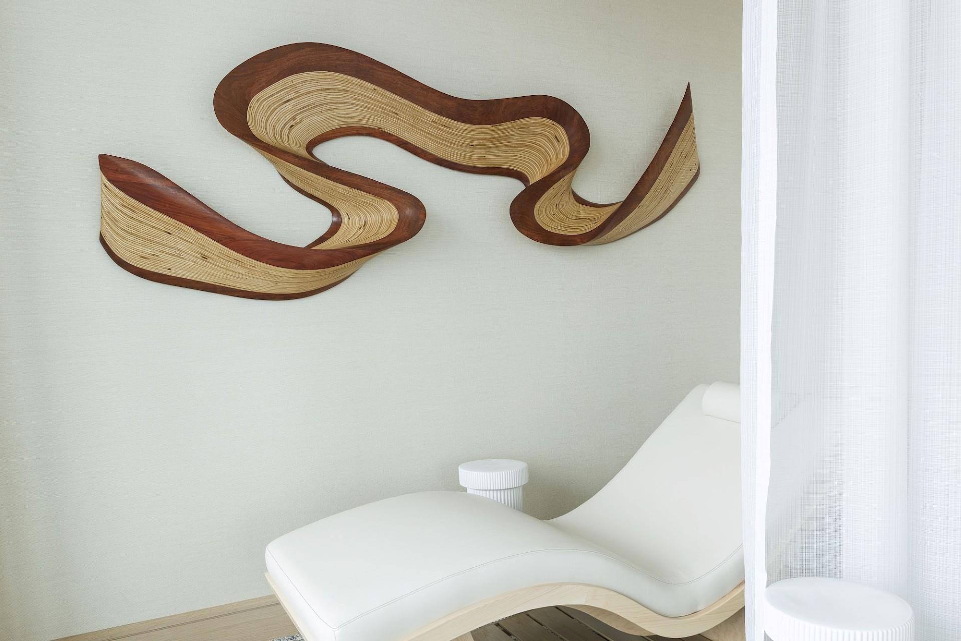 Spa relaxation area with a white contoured lounge chair beneath an abstract layered wood wall sculpture and sheer privacy curtain.