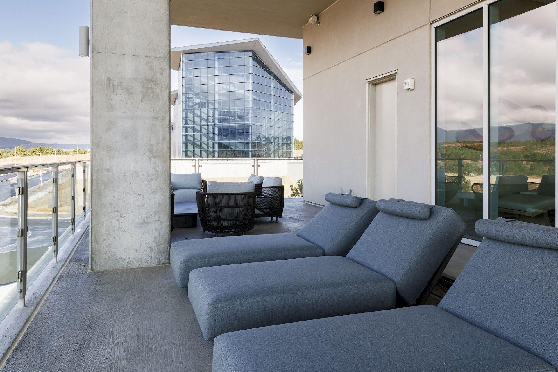 Outdoor terrace with cushioned gray lounge chairs, glass railings, and views of a modern glass building and distant mountains.