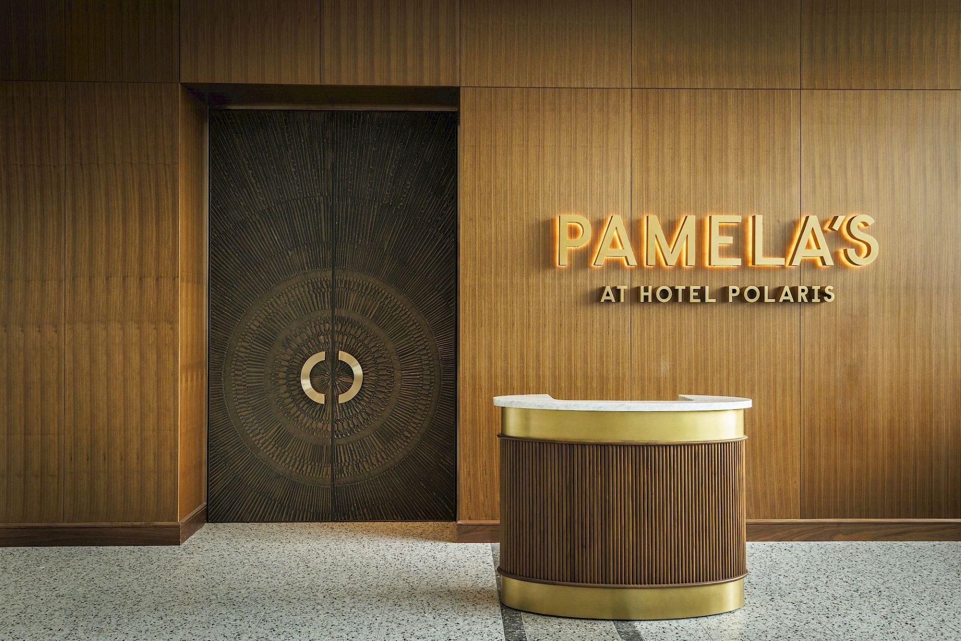 Entrance to Pamela’s at Hotel Polaris with illuminated gold signage, wood-paneled walls, decorative dark double doors, and a curved host stand.