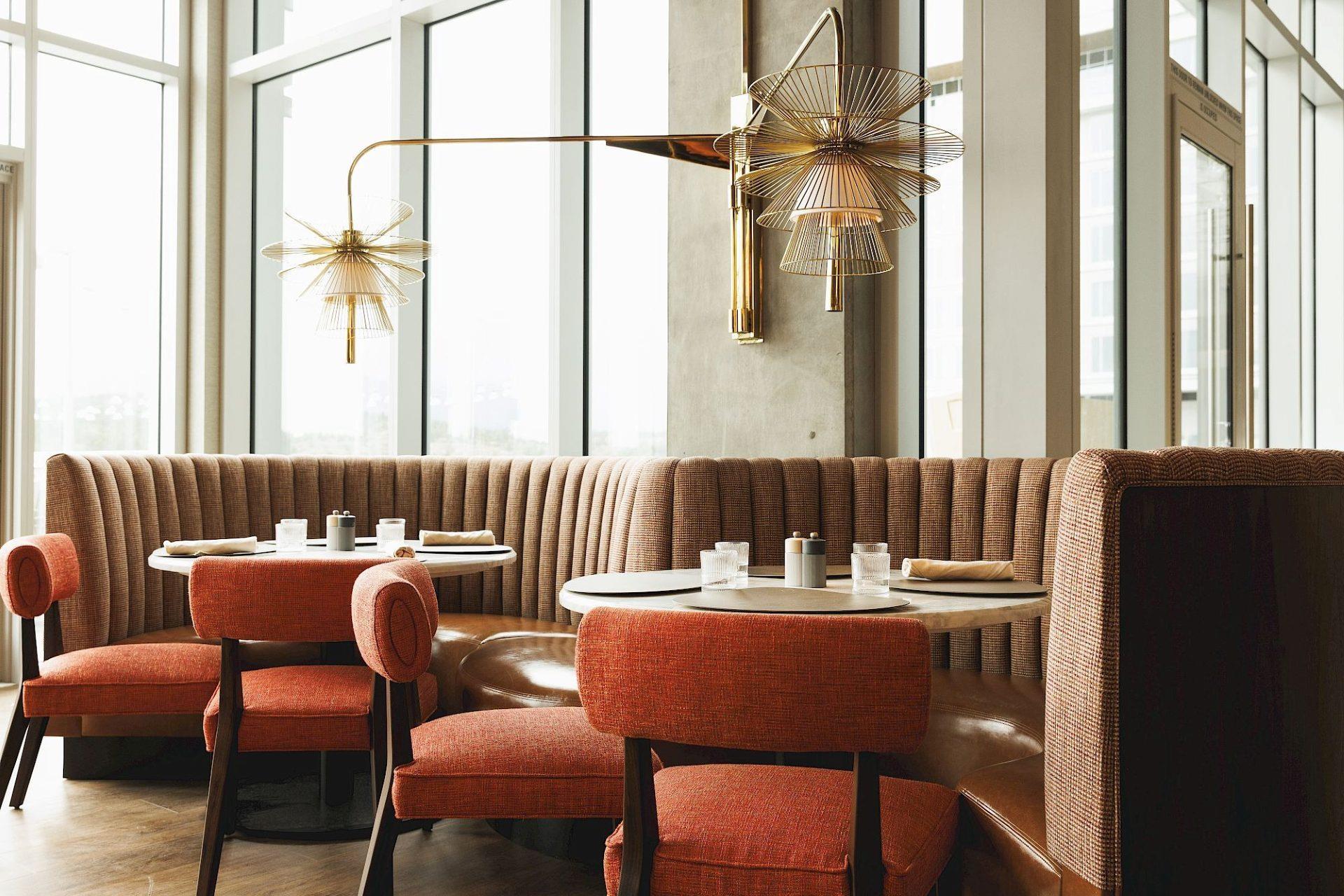 Restaurant seating area with curved brown banquettes, coral upholstered chairs, round tables, sculptural brass wall lights, and floor-to-ceiling windows.