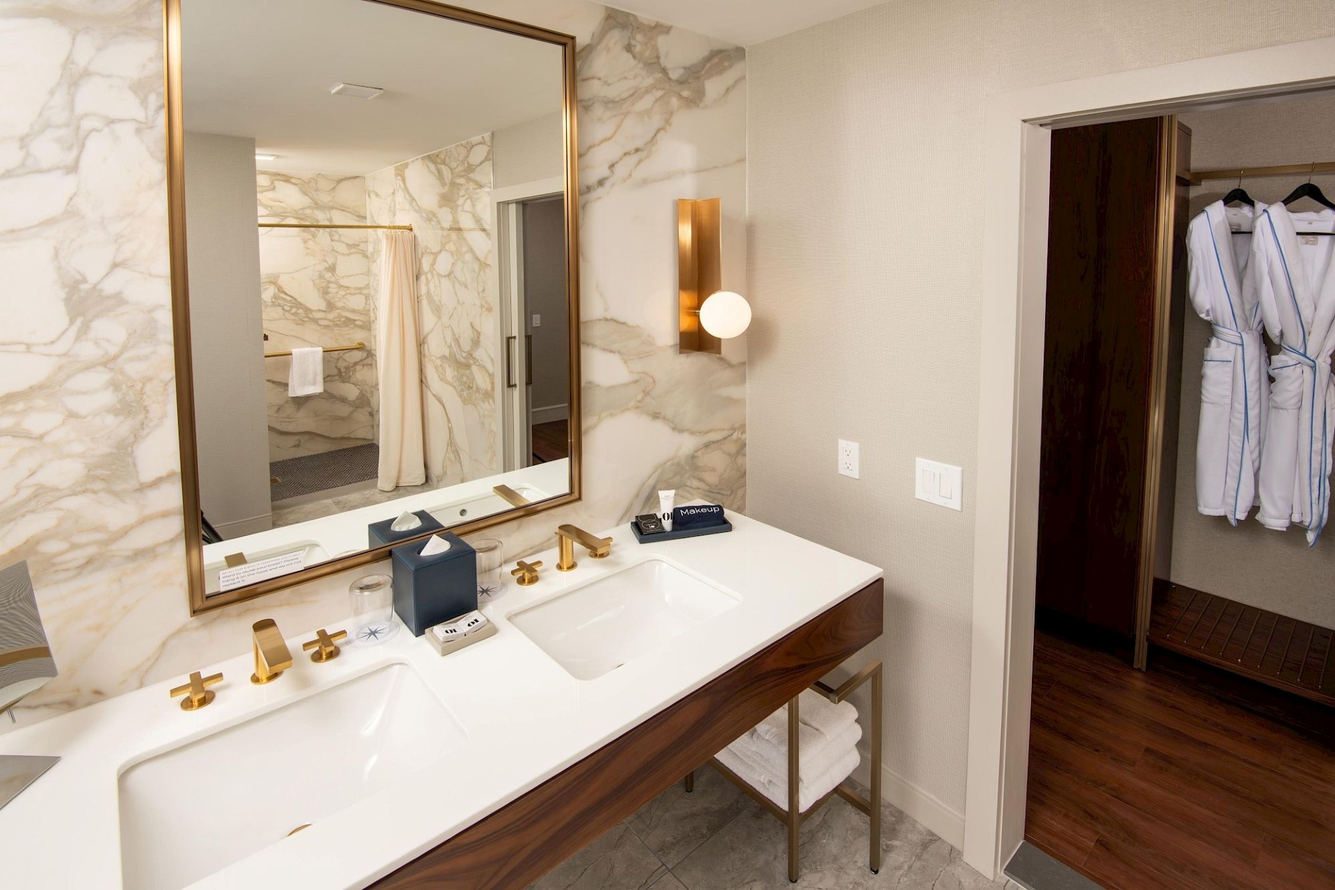 Accessible suite bathroom with double-sink white vanity, wood cabinetry, brass fixtures, large framed mirror, marble-patterned walls, and adjacent open closet with robes.