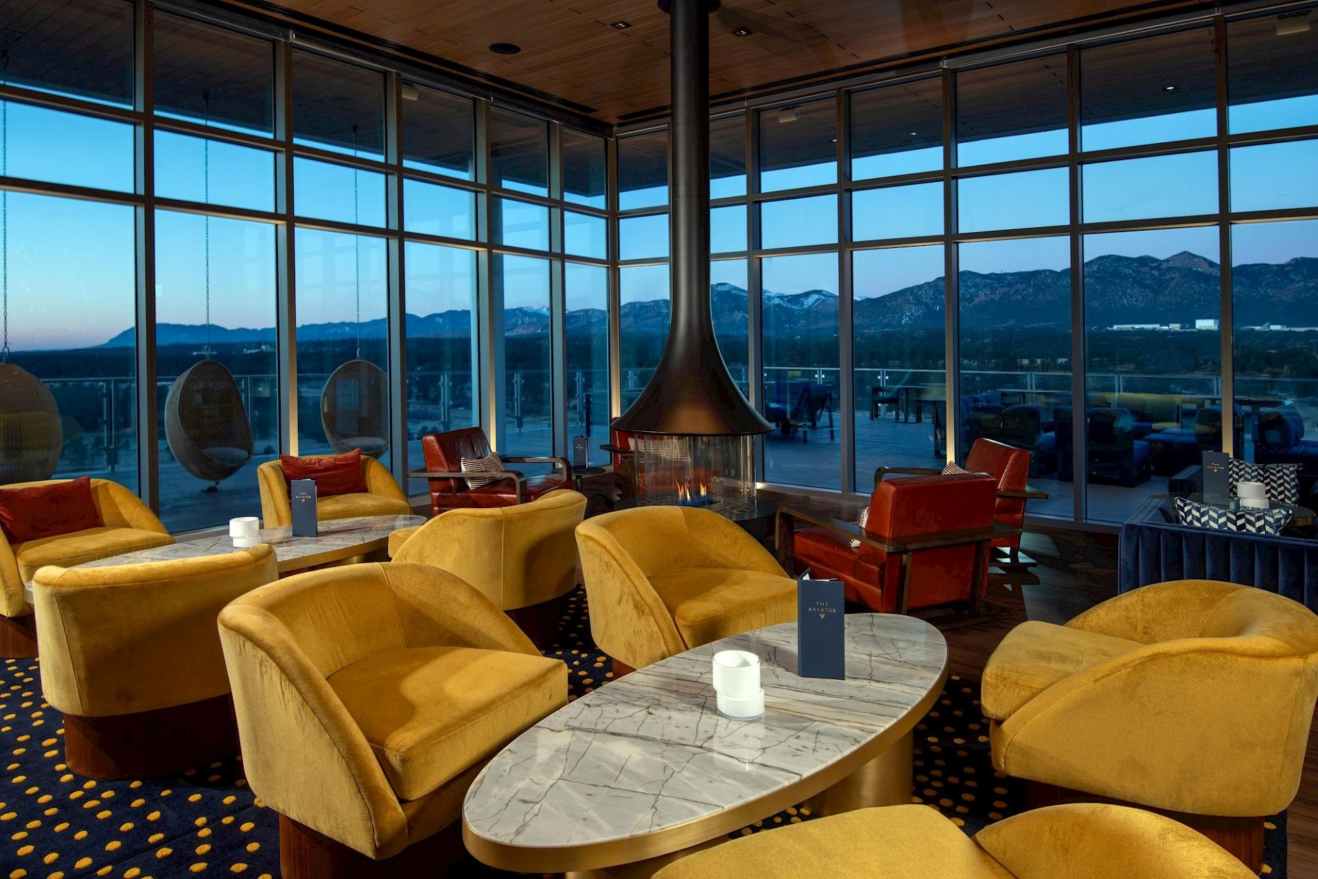 Upscale lounge with mustard-yellow chairs, red leather seating, marble tables, a central fireplace, and floor-to-ceiling windows overlooking mountains at dusk.