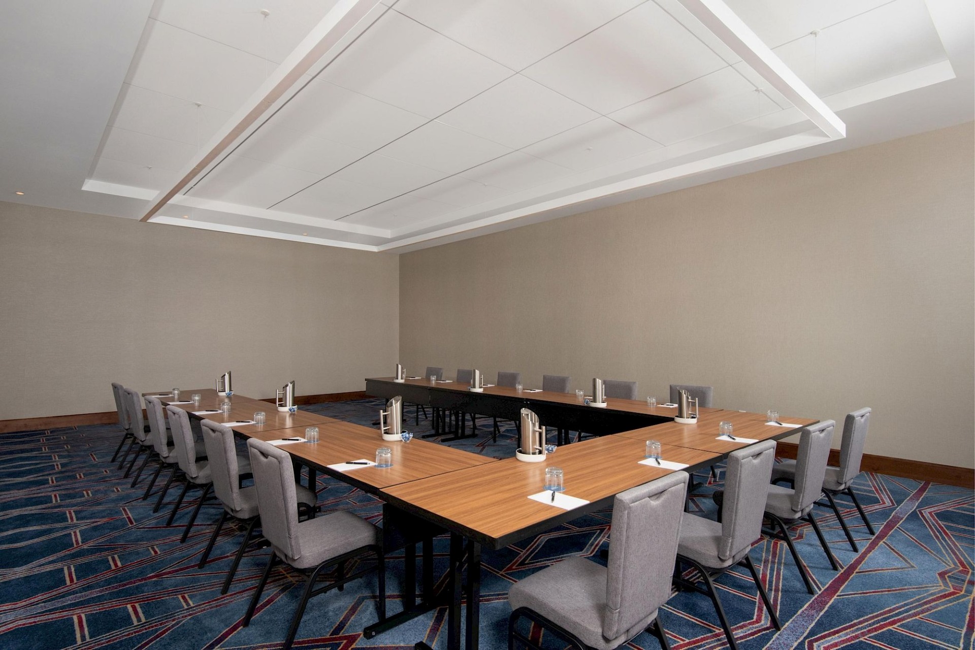 Modern meeting room with wood tables arranged in a U-shape, gray upholstered chairs, water service, and geometric blue patterned carpet.