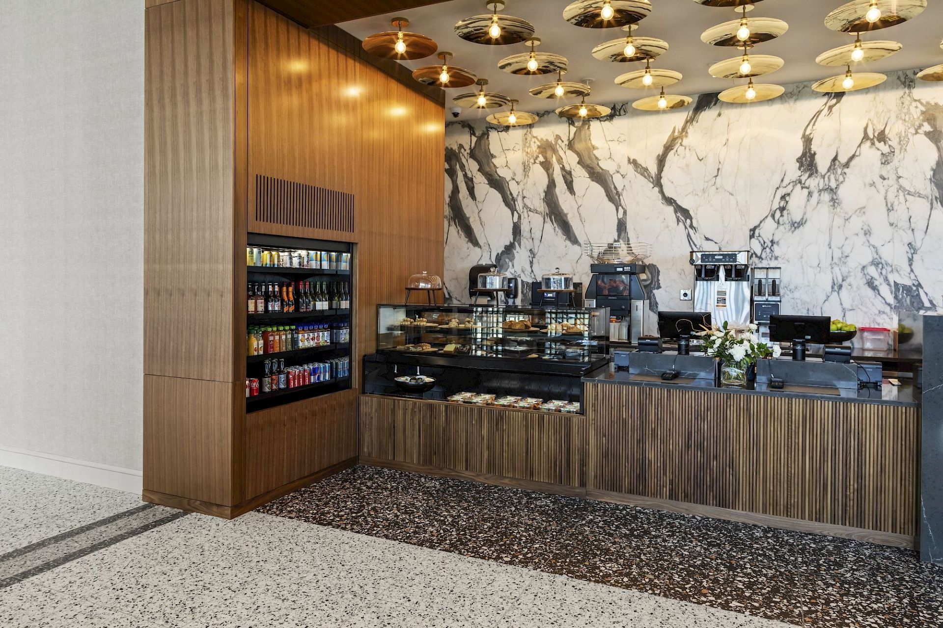 Modern grab-and-go café counter with wood cabinetry, refrigerated drinks, pastry displays, coffee machines, marble-patterned wall, and decorative ceiling lights.