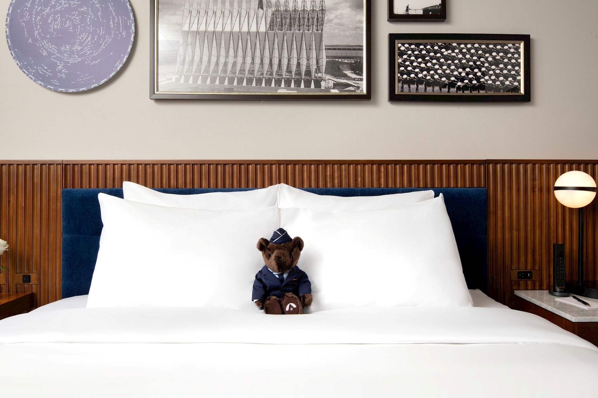 Close view of a king bed with crisp white pillows, blue upholstered headboard, wood slat paneling, globe bedside lamps, framed artwork, and a uniformed teddy bear.