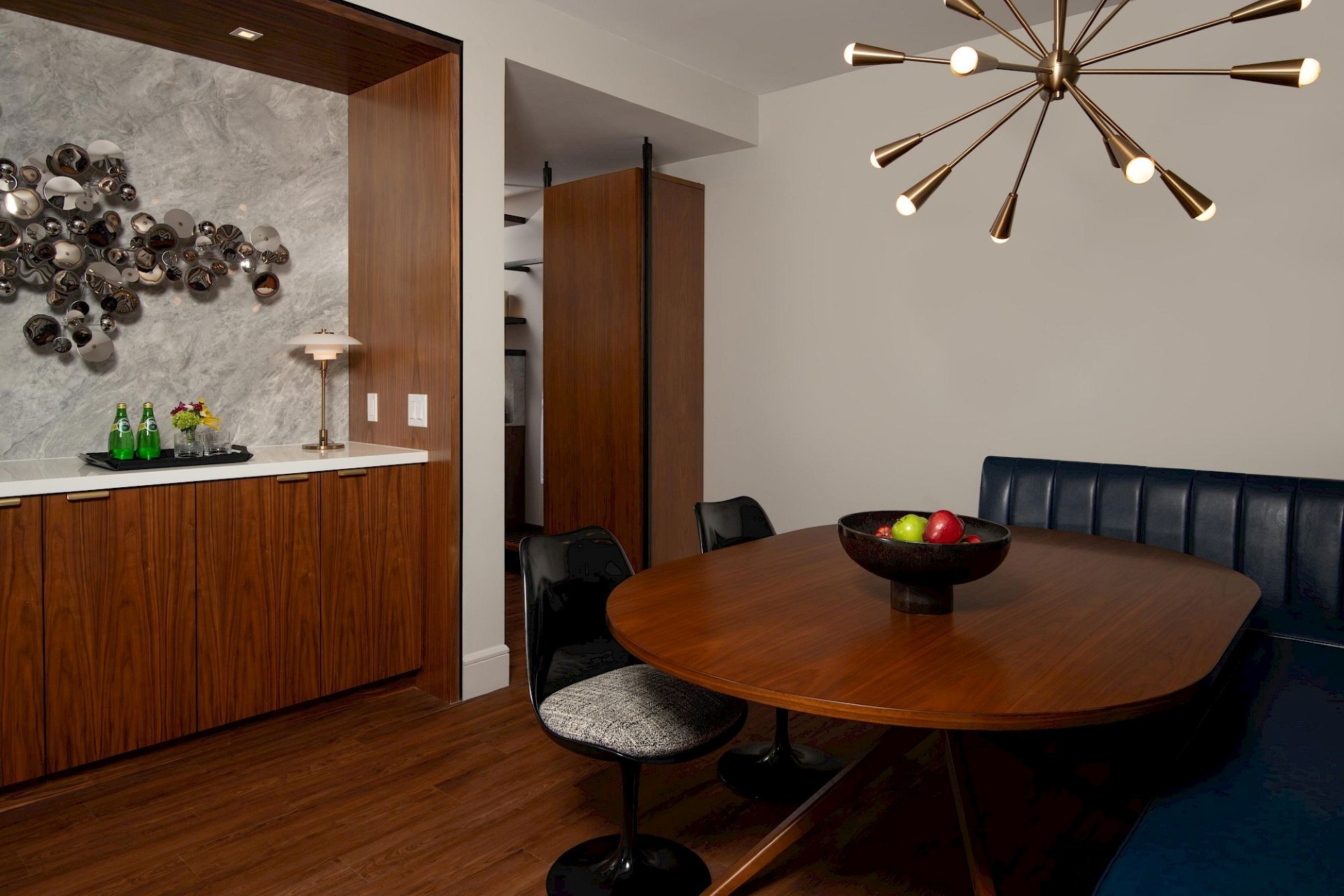 Suite dining area with rounded wood table, navy banquette, black chairs, starburst brass chandelier, and adjacent wood wet bar with sculptural wall art.