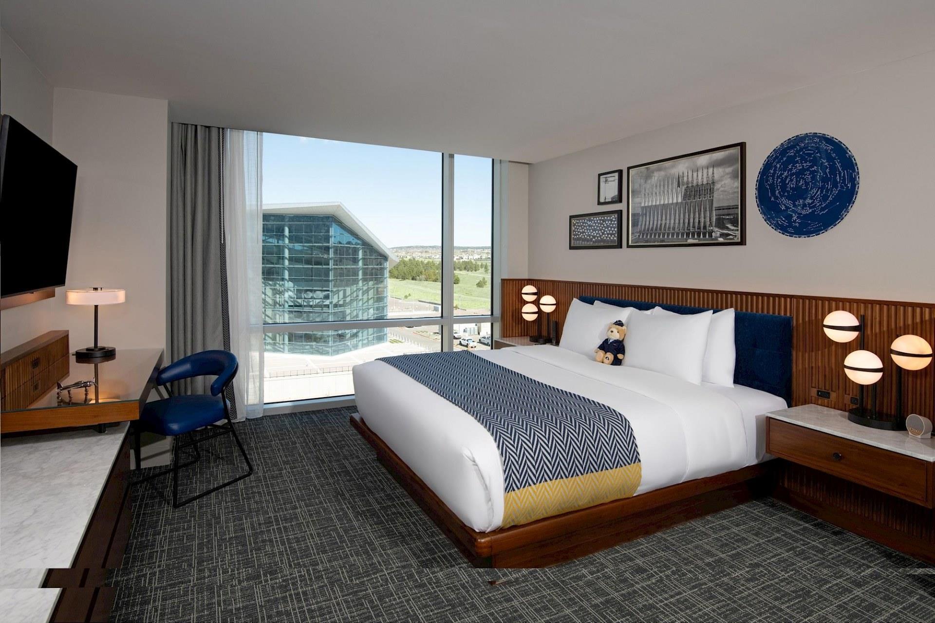 Mountain-view suite bedroom with king bed, white bedding, blue patterned throw, full-height wood wall, cream chaise, desk, and expansive window.