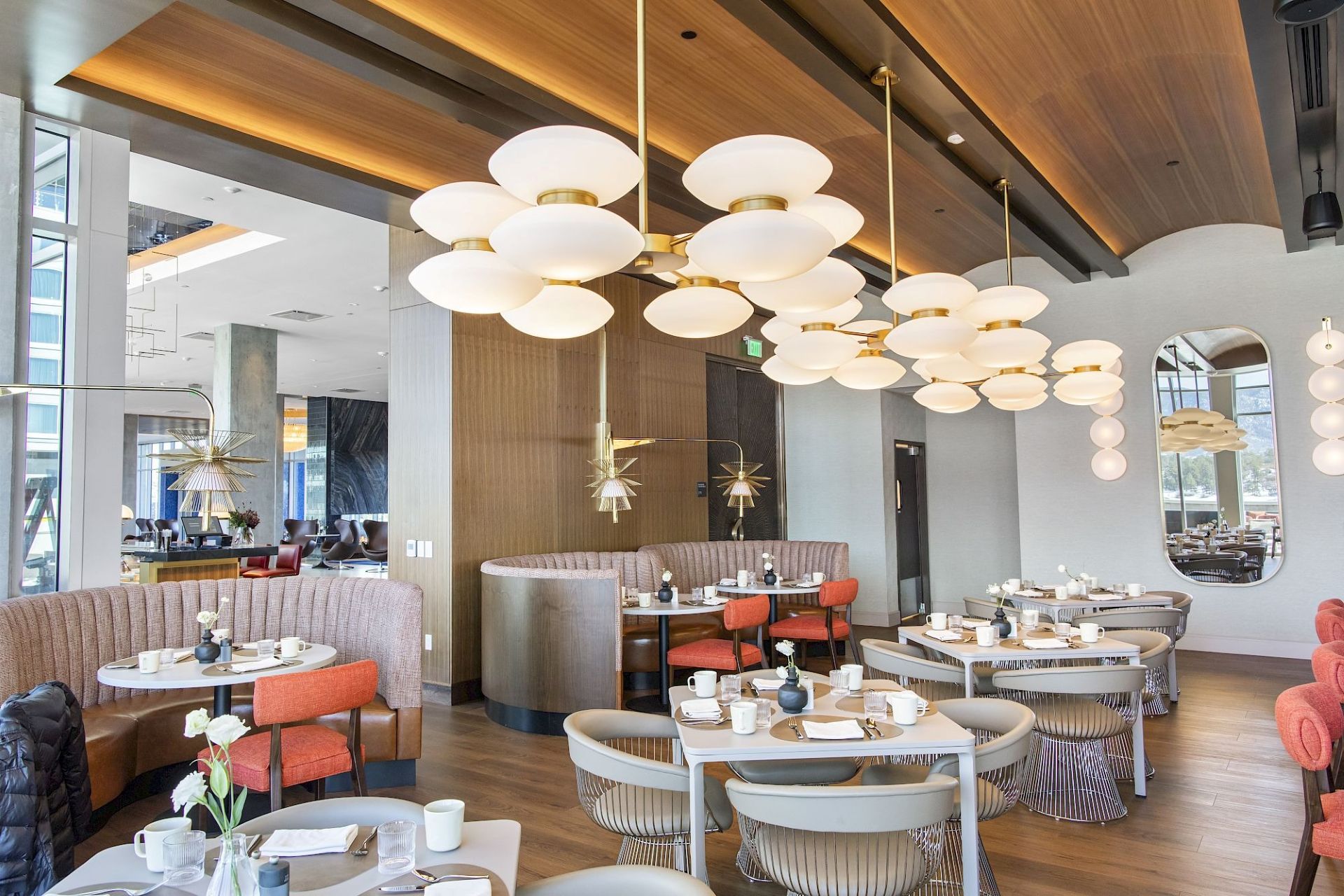 Bright restaurant dining room with curved booths, coral chairs, wood-paneled ceiling, sculptural globe chandeliers, and floor-to-ceiling windows.