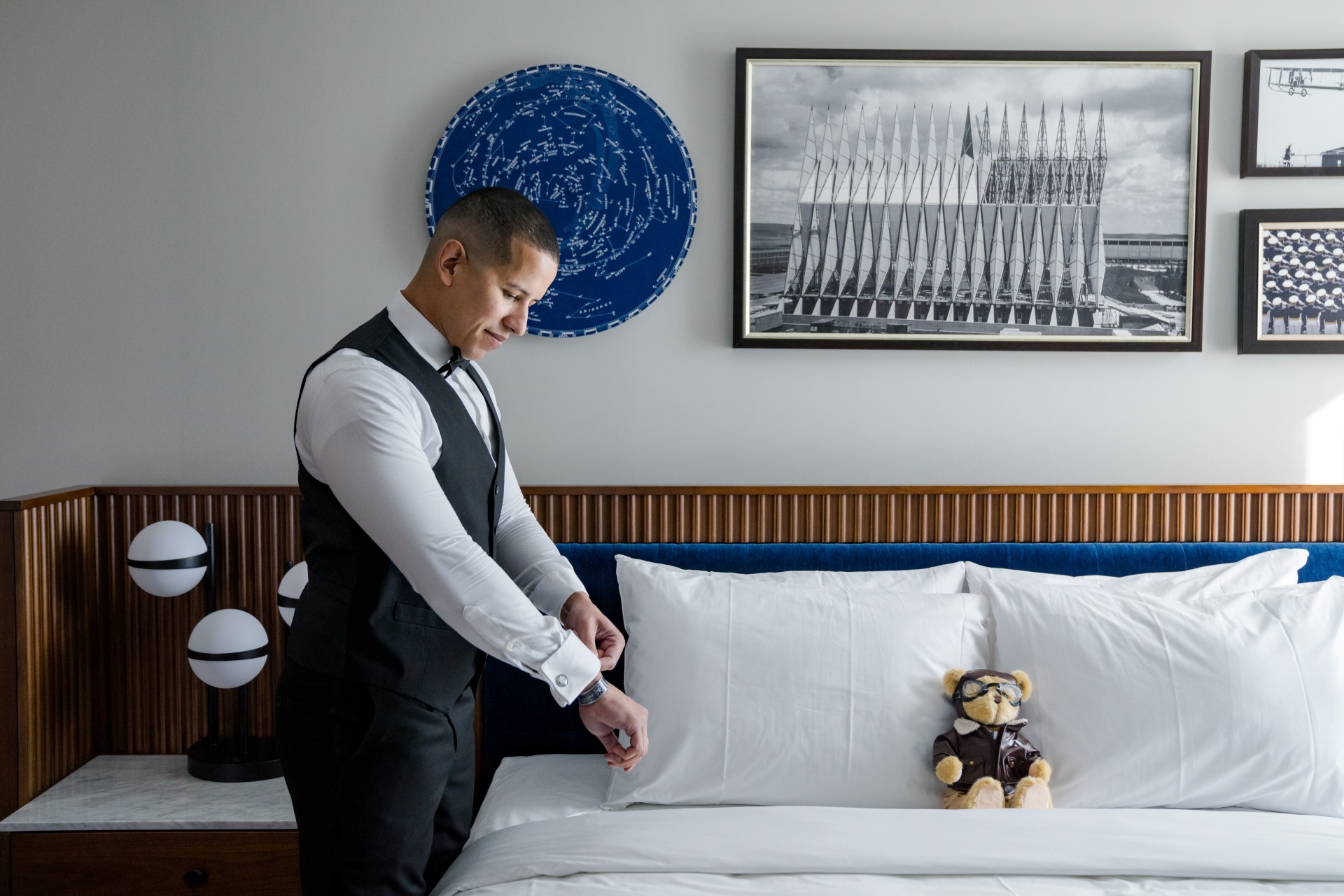 Groom adjusts his cufflinks beside a bed in a Hotel Polaris guest room, with aviation-inspired artwork and a pilot teddy bear.