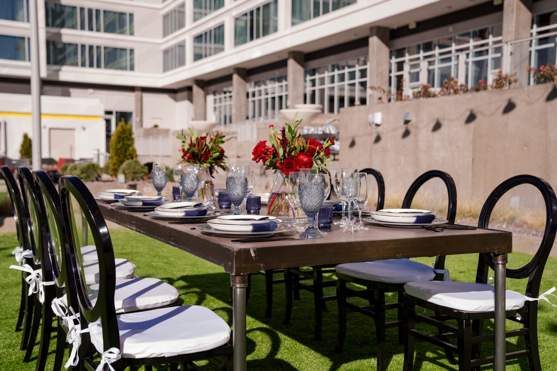 Outdoor table set for a meal with eight black chairs, white cushions, glassware, plates, and red floral centerpieces in front of a modern building.