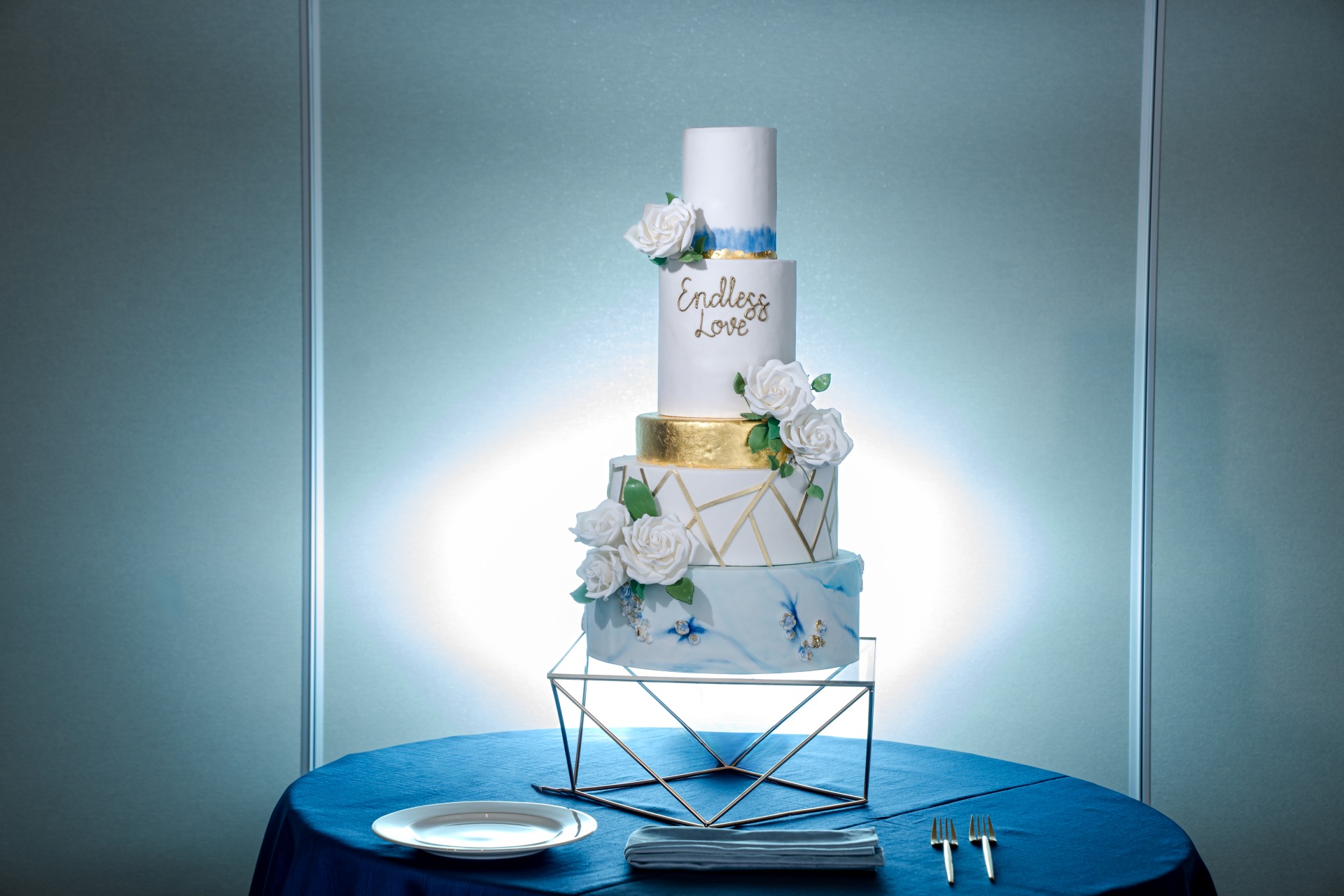A four-tiered wedding cake with white roses and geometric gold accents sits on a blue tablecloth, with plates and forks beside it.