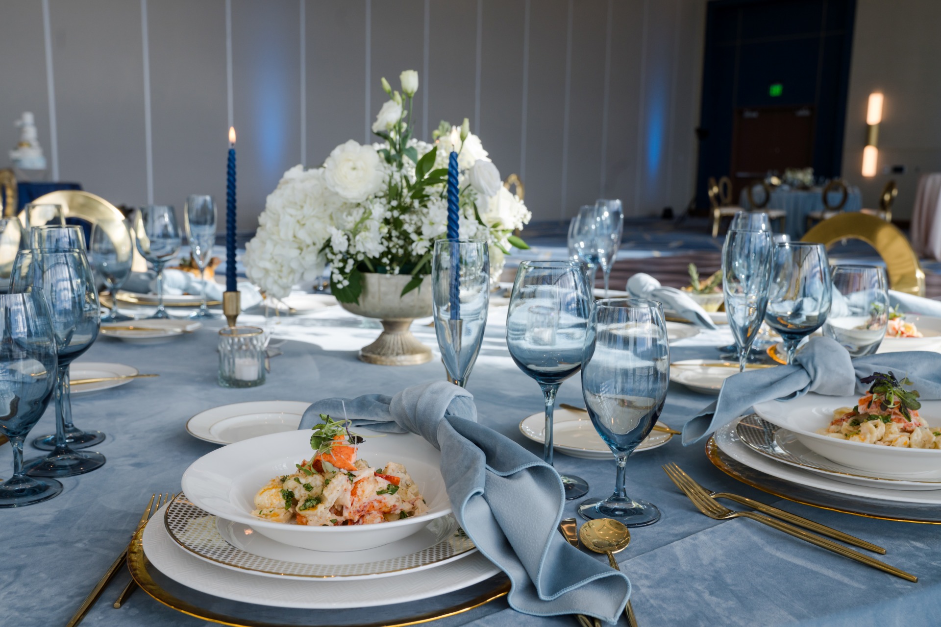 Elegant banquet table set for a formal event with blue linens, gold-accented place settings, white floral centerpieces, blue glassware, and plated seafood dishes in Hotel Polaris' event space.