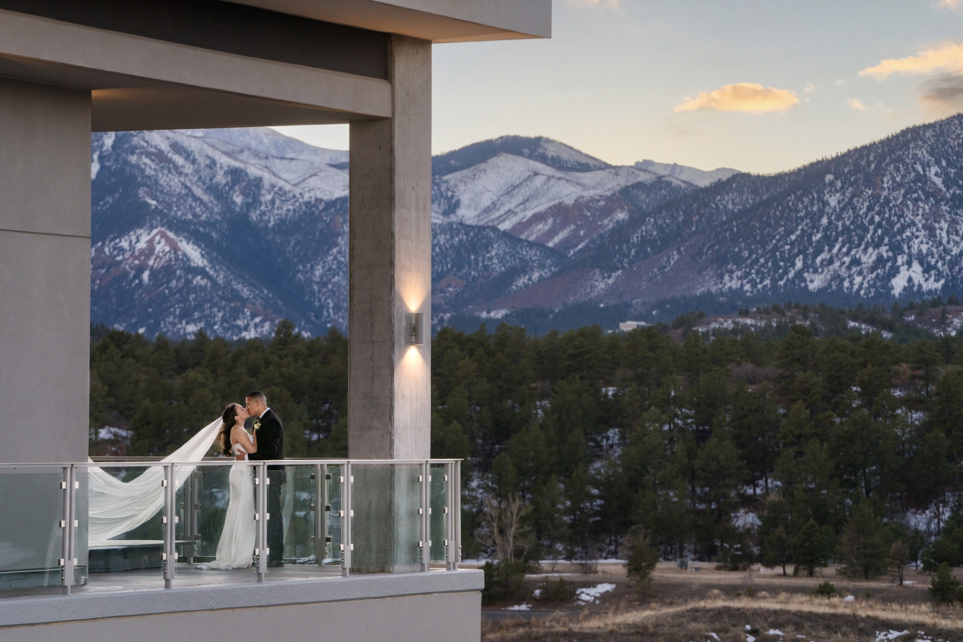 A bride and groom stand on a balcony at sunset, overlooking a forest and snow-capped mountains in the background.