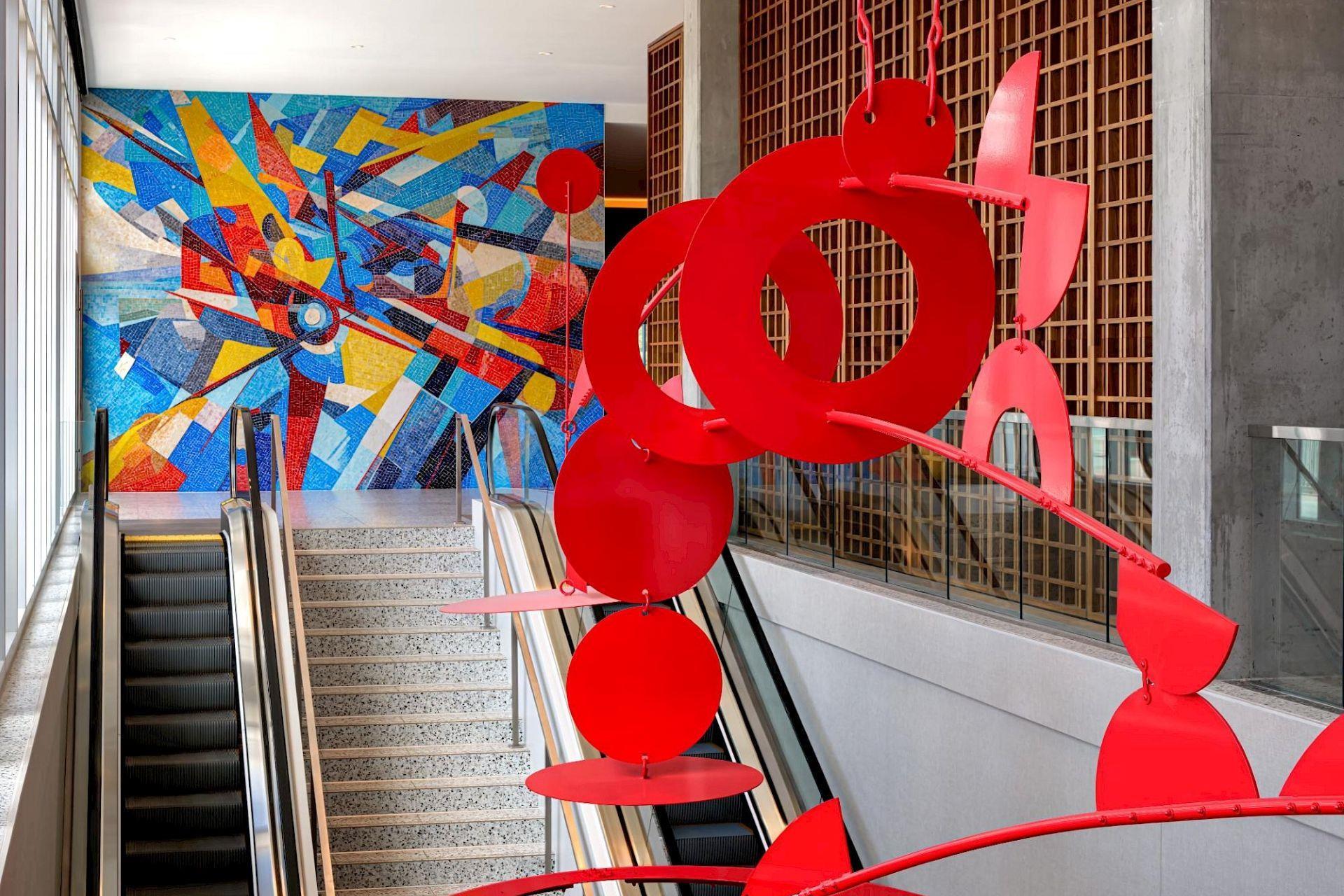 Red suspended sculpture above escalators at Hotel Polaris, with a colorful aviation-inspired mosaic in the background.