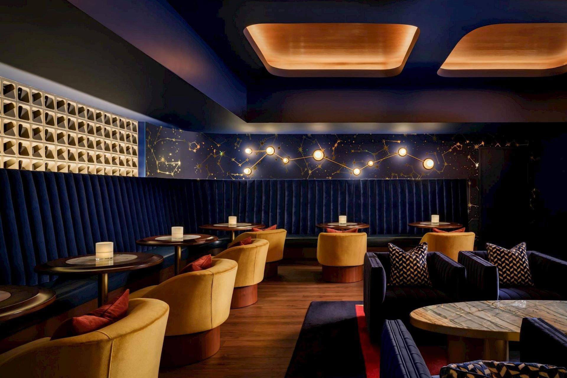 Intimate lounge with deep blue banquette seating, mustard chairs, constellation-inspired wall art and lighting, wood floors, and marble-topped tables.
