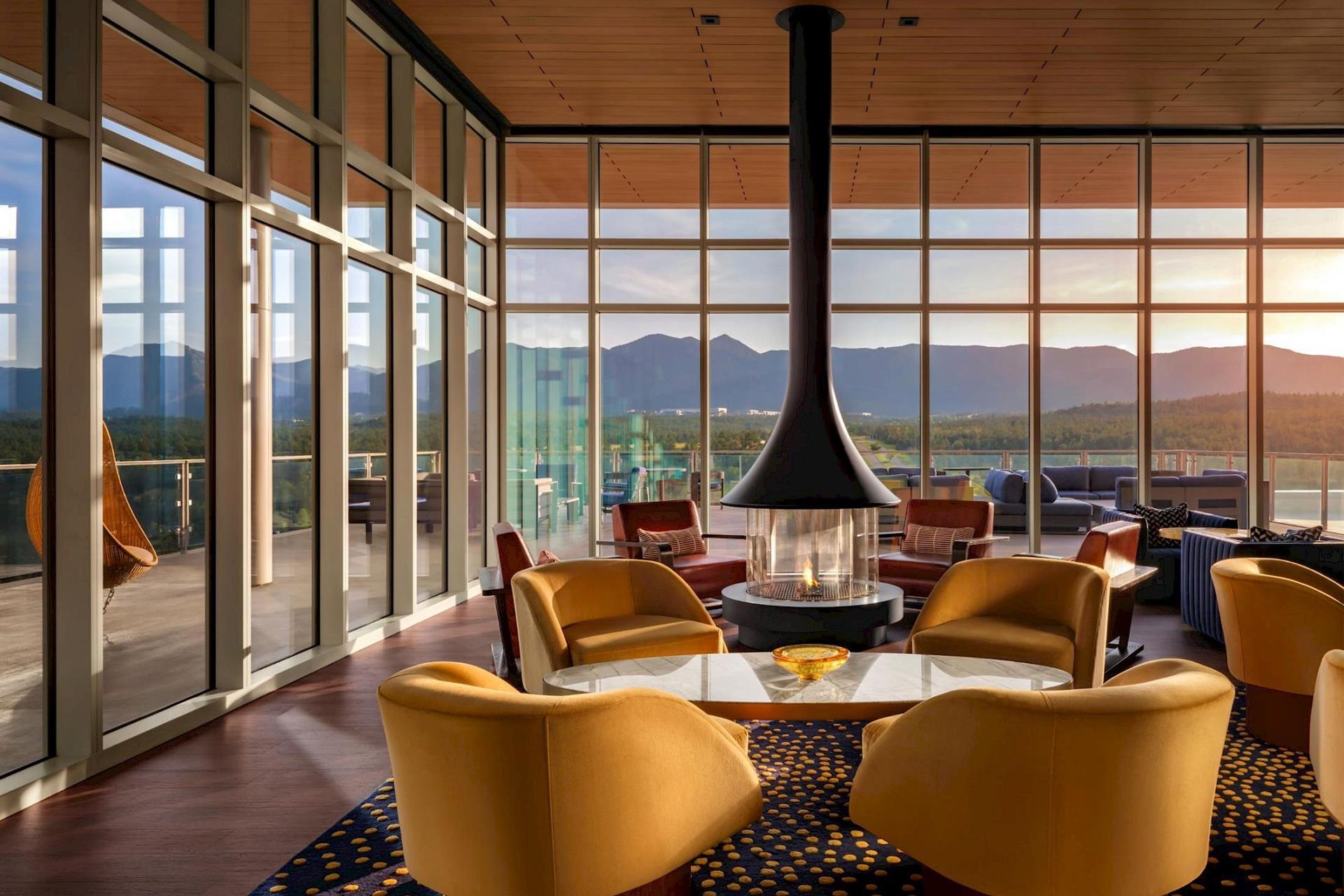 Rooftop lounge at Hotel Polaris featuring a suspended fireplace, contemporary seating, floor-to-ceiling windows, and panoramic views of the Rocky Mountains at sunset.