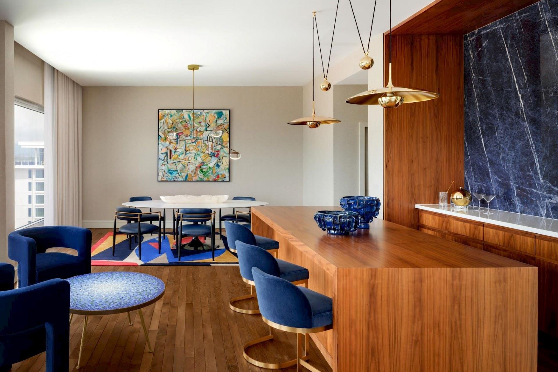 Contemporary suite living and dining area with large wood bar, blue velvet seating, brass pendant lights, colorful abstract artwork, and dark marble accent wall.