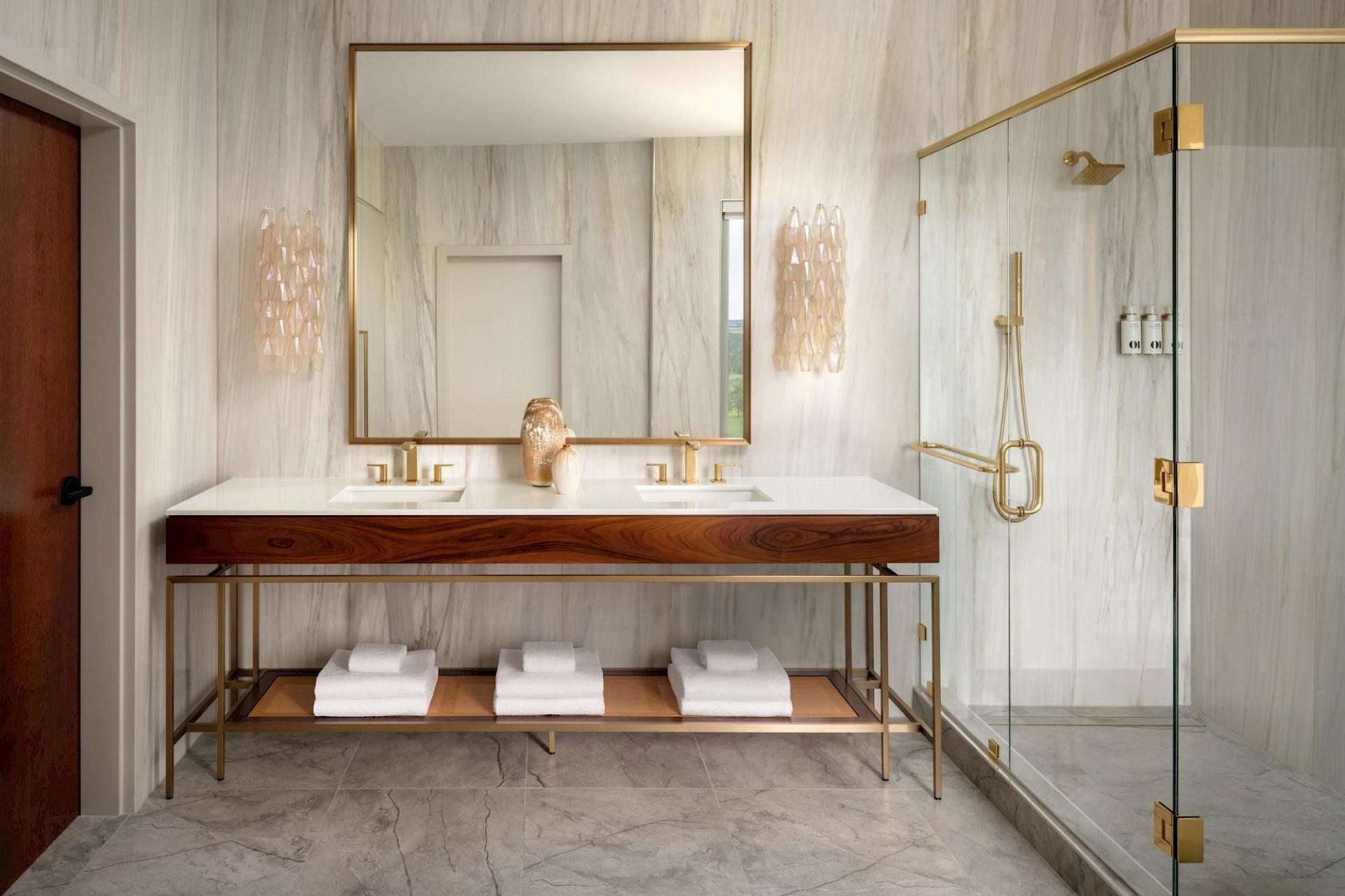 Spacious bathroom with double wood vanity, white countertop, brass-framed mirror, decorative sconces, and glass-enclosed shower with brass hardware.