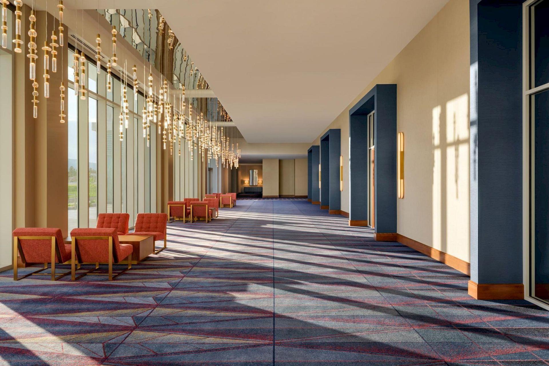 Sunlit hotel corridor with floor-to-ceiling windows, cascading pendant lights, coral lounge chairs, blue architectural accents, and patterned carpet.