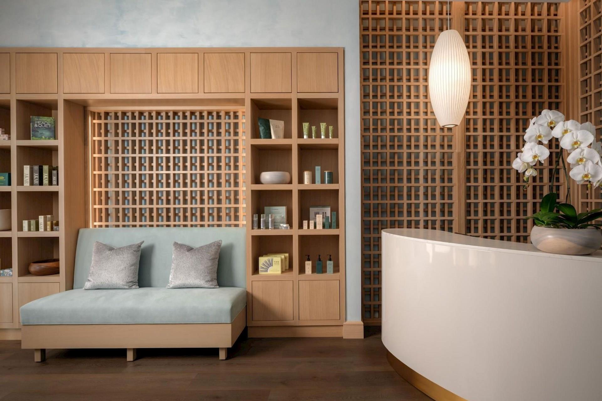 Serene spa reception area with light wood shelving, a pale blue bench, lattice wood screens, curved white desk, pendant light, and white orchids.