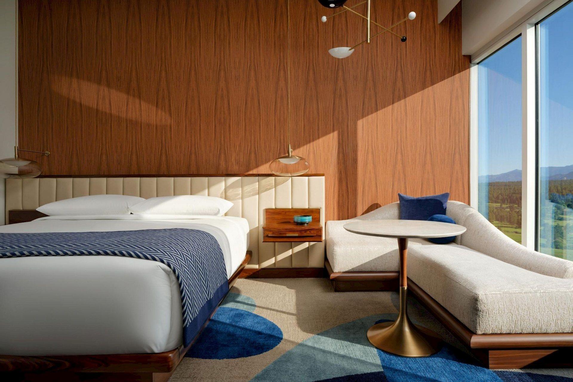 Modern king guest room with warm wood-paneled walls, white bedding, blue patterned throw, cream chaise lounge, and floor-to-ceiling mountain views.