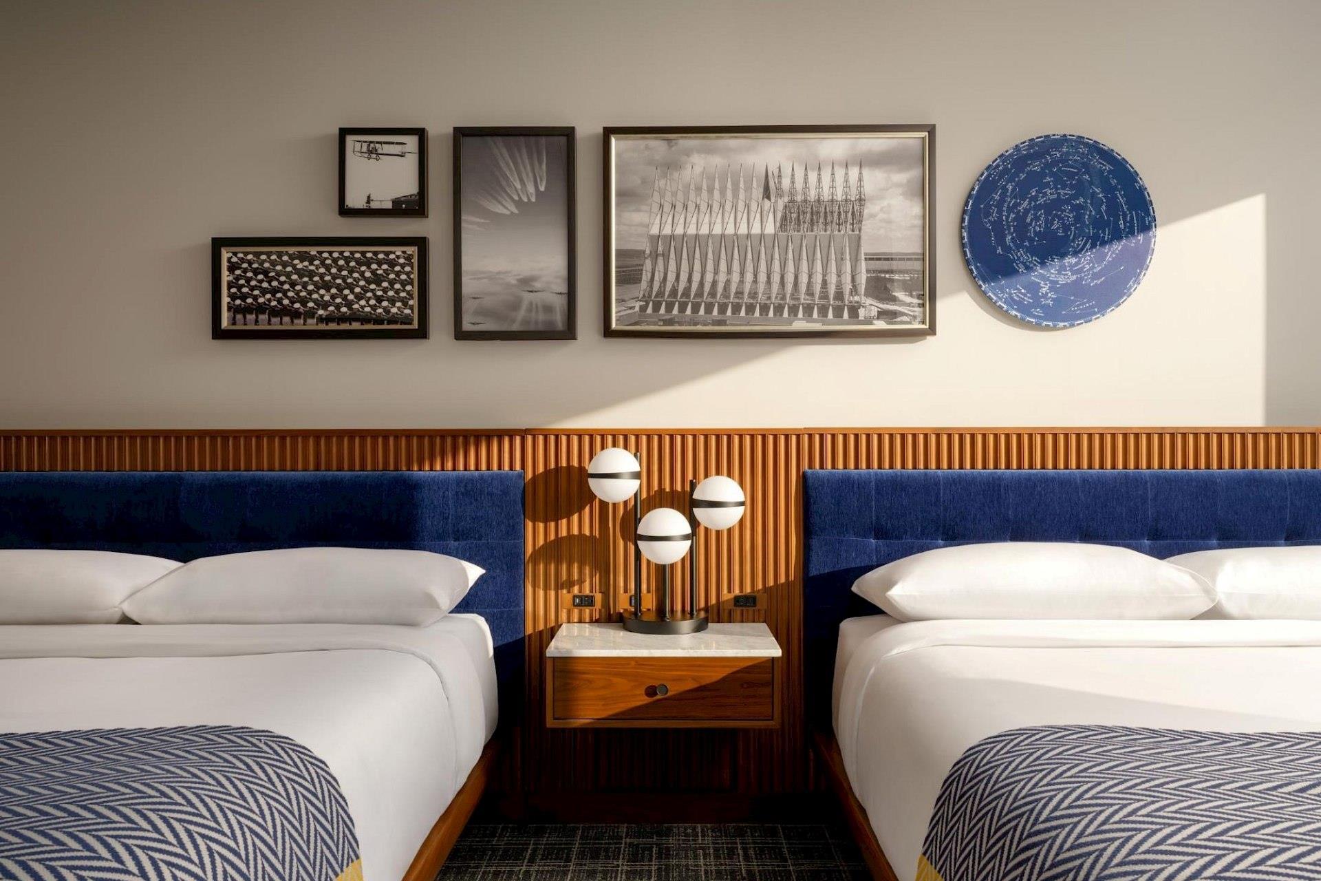 Double guest room with two white beds, deep blue upholstered headboards, wood slat wall paneling, framed black-and-white artwork, and a central globe lamp.