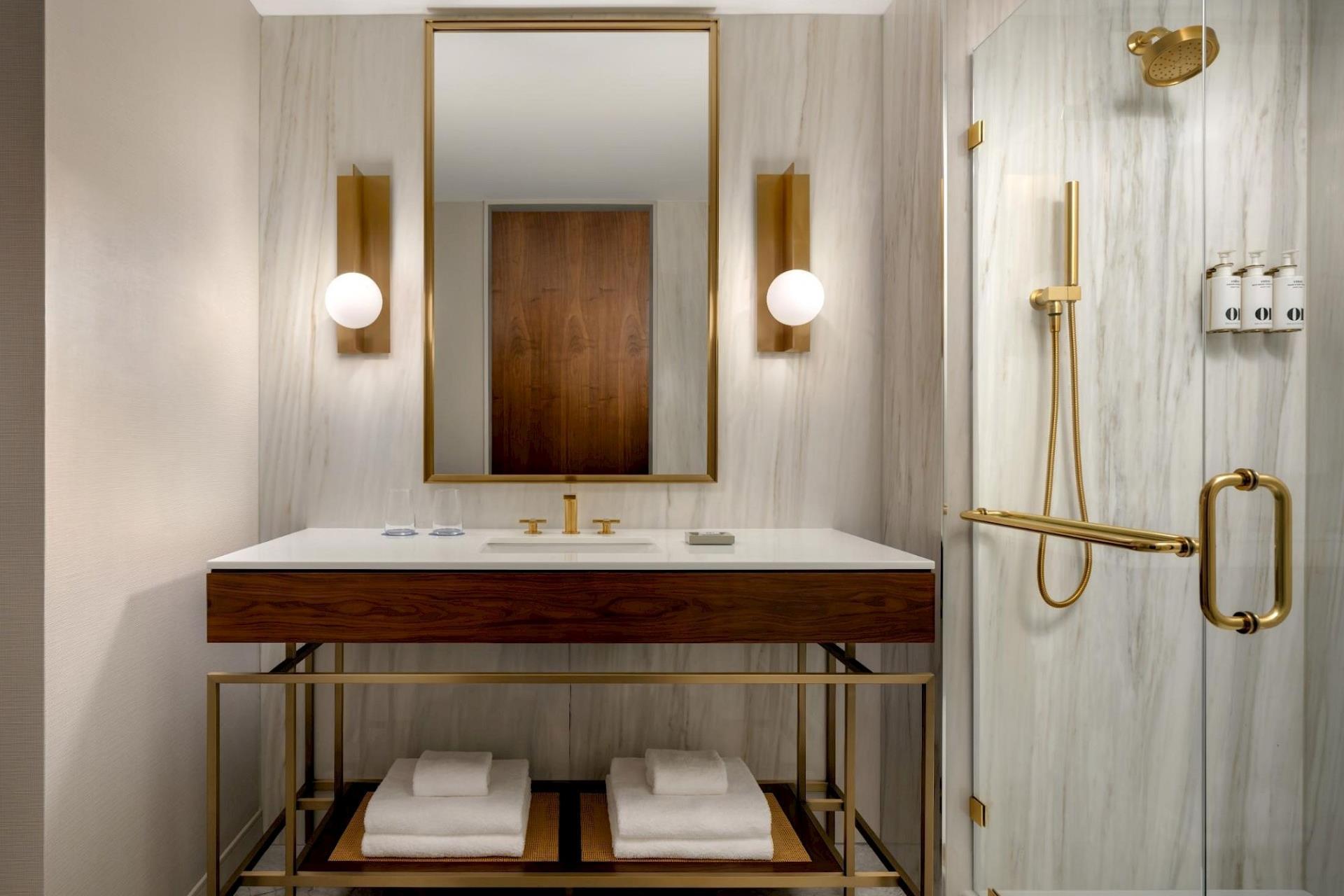 Bright hotel bathroom with wood vanity, white countertop, large brass-framed mirror, globe sconces, and glass shower with brass fixtures.