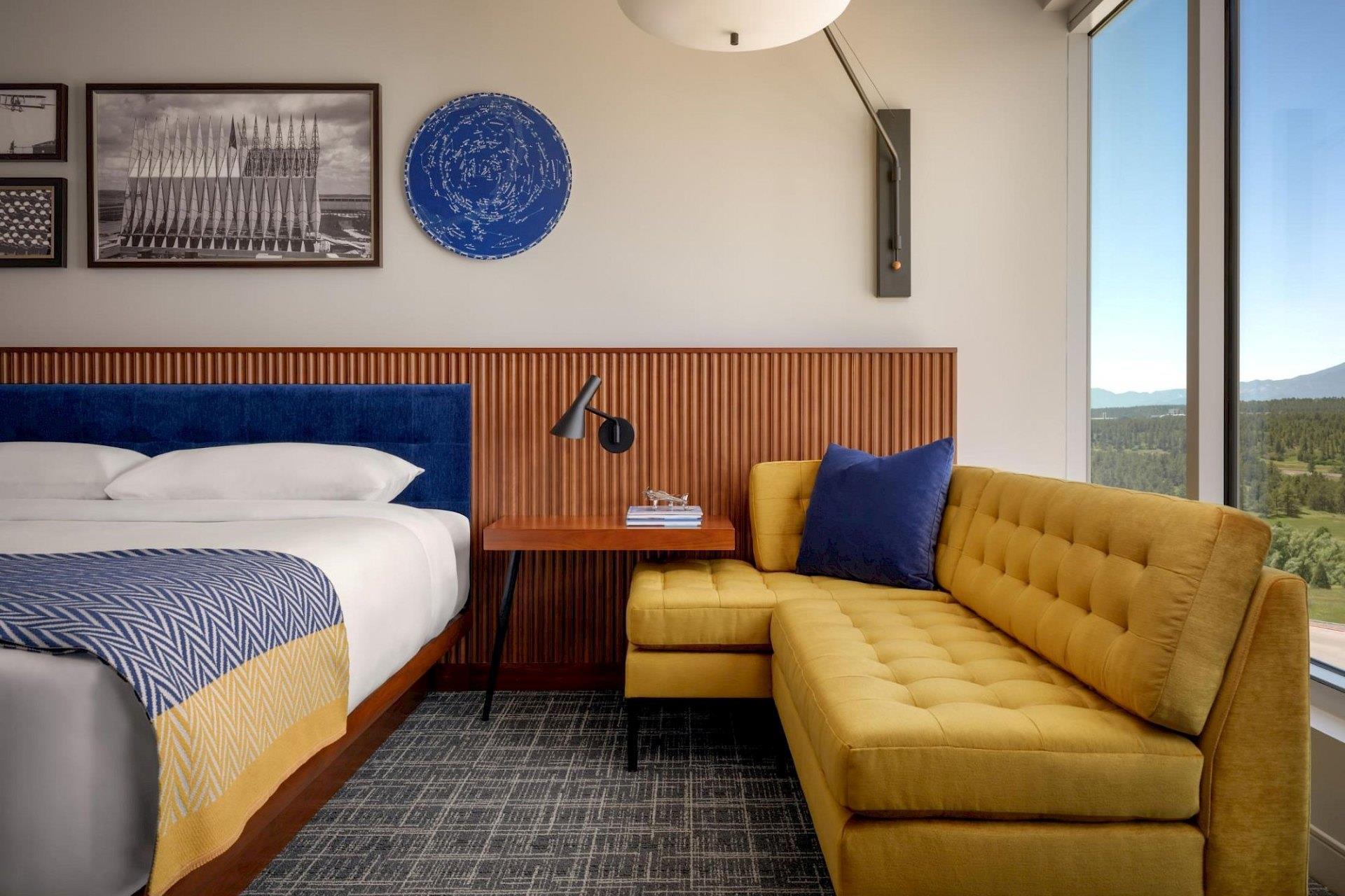 Guest room seating area with mustard-yellow sectional sofa, navy pillow, wood-paneled wall, blue headboard, and large window overlooking mountains.