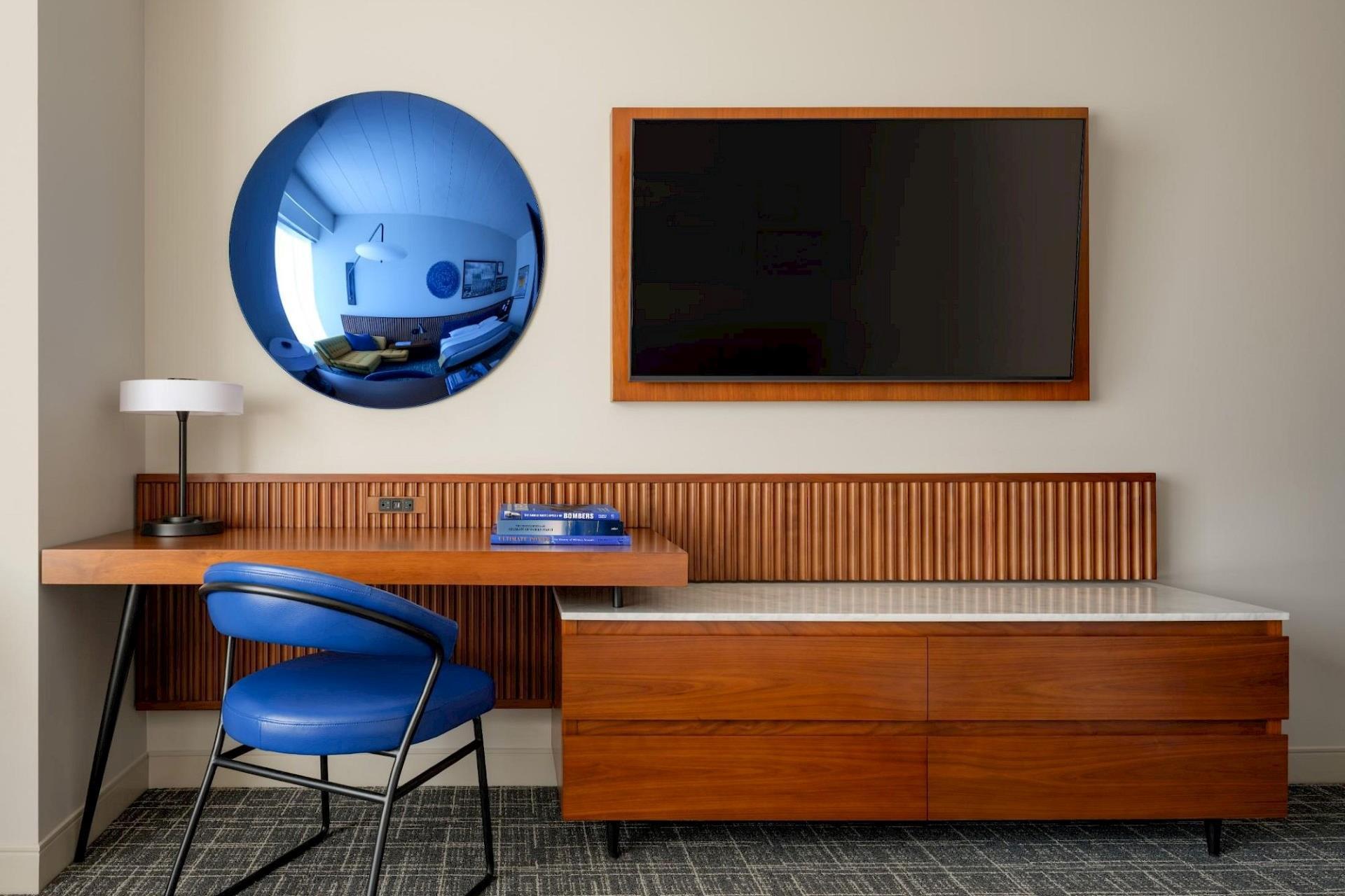 Guest room workspace with blue upholstered chair, floating wood desk, round blue-tinted mirror, wall-mounted TV, and low wood dresser.