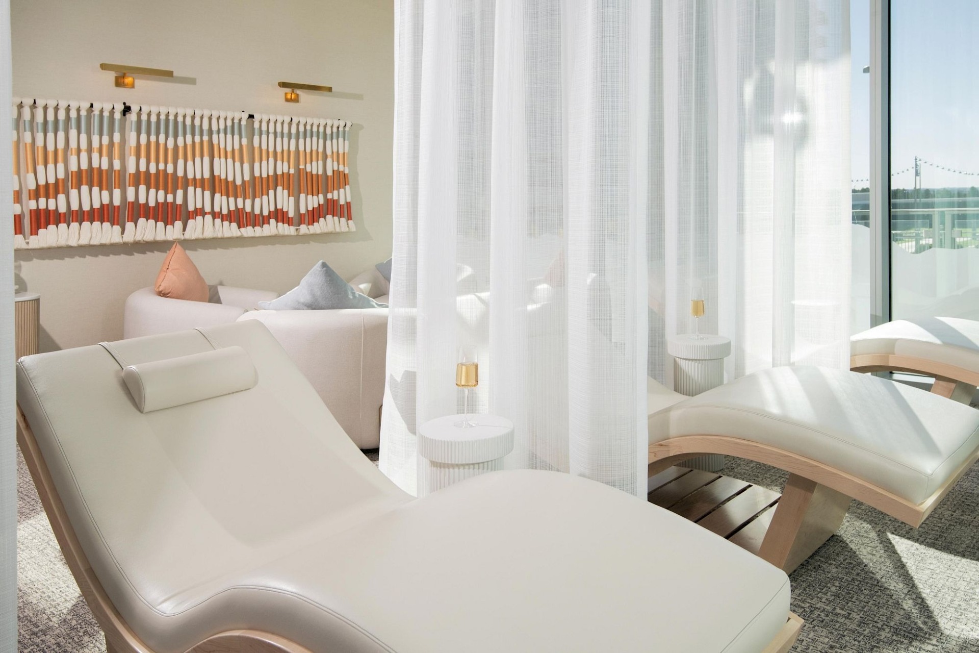 Sunlit spa relaxation room with white contoured lounge chairs, sheer privacy curtains, small side tables, and colorful woven wall art.