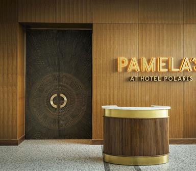 Entrance to Pamela’s at Hotel Polaris with illuminated gold signage, wood-paneled walls, decorative dark double doors, and a curved host stand.