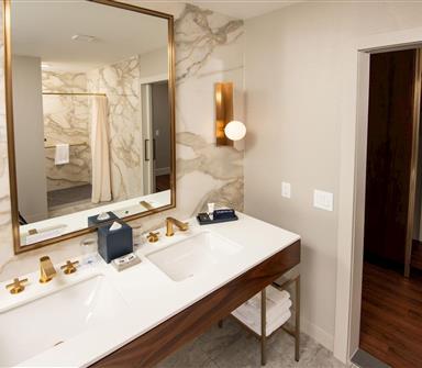 Accessible suite bathroom with double-sink white vanity, wood cabinetry, brass fixtures, large framed mirror, marble-patterned walls, and adjacent open closet with robes.
