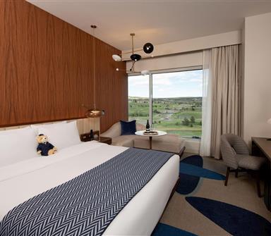 Club-level king room with white bedding, blue headboard, patterned throw, wood slat paneling, blue desk chair, framed artwork, and floor-to-ceiling window.