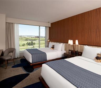 Club-level room with two white beds, blue patterned throws, warm wood accent wall, large window, desk, and neutral upholstered chair.