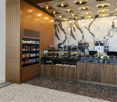 Modern grab-and-go café counter with wood cabinetry, refrigerated drinks, pastry displays, coffee machines, marble-patterned wall, and decorative ceiling lights.