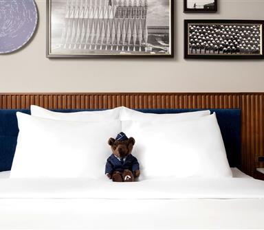 Close view of a king bed with crisp white pillows, blue upholstered headboard, wood slat paneling, globe bedside lamps, framed artwork, and a uniformed teddy bear.