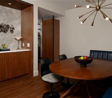 Suite dining area with rounded wood table, navy banquette, black chairs, starburst brass chandelier, and adjacent wood wet bar with sculptural wall art.