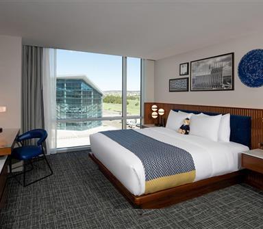 Mountain-view suite bedroom with king bed, white bedding, blue patterned throw, full-height wood wall, cream chaise, desk, and expansive window.