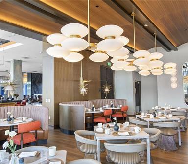 Bright restaurant dining room with curved booths, coral chairs, wood-paneled ceiling, sculptural globe chandeliers, and floor-to-ceiling windows.