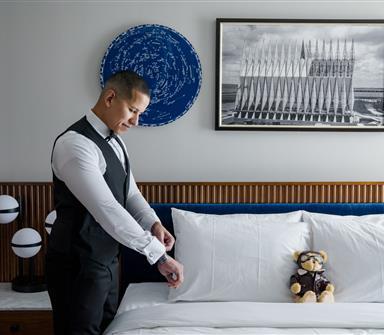 Groom adjusts his cufflinks beside a bed in a Hotel Polaris guest room, with aviation-inspired artwork and a pilot teddy bear.