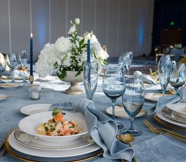 Elegant banquet table set for a formal event with blue linens, gold-accented place settings, white floral centerpieces, blue glassware, and plated seafood dishes in Hotel Polaris' event space.