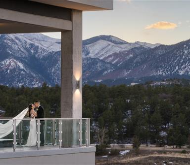 A bride and groom stand on a balcony at sunset, overlooking a forest and snow-capped mountains in the background.