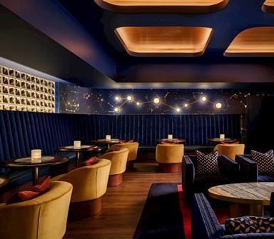 Intimate lounge with deep blue banquette seating, mustard chairs, constellation-inspired wall art and lighting, wood floors, and marble-topped tables.