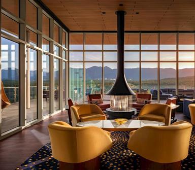 Rooftop lounge at Hotel Polaris featuring a suspended fireplace, contemporary seating, floor-to-ceiling windows, and panoramic views of the Rocky Mountains at sunset.