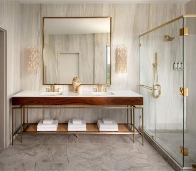 Spacious bathroom with double wood vanity, white countertop, brass-framed mirror, decorative sconces, and glass-enclosed shower with brass hardware.