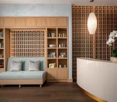 Serene spa reception area with light wood shelving, a pale blue bench, lattice wood screens, curved white desk, pendant light, and white orchids.
