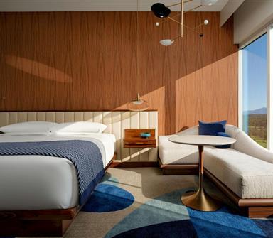 Modern king guest room with warm wood-paneled walls, white bedding, blue patterned throw, cream chaise lounge, and floor-to-ceiling mountain views.