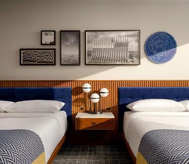 Double guest room with two white beds, deep blue upholstered headboards, wood slat wall paneling, framed black-and-white artwork, and a central globe lamp.