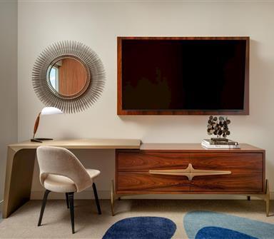 Guest room work area with a curved desk, upholstered chair, sunburst mirror, wall-mounted TV, and mid-century wood dresser with brass detailing.